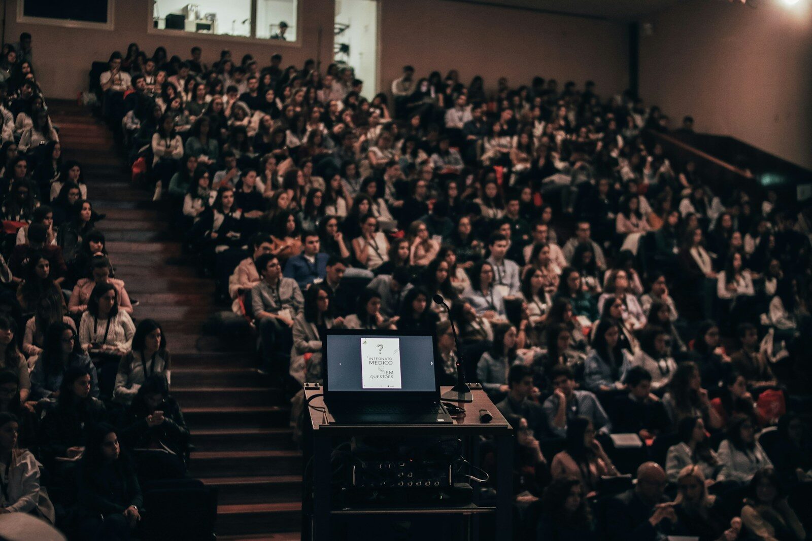 A large audience attentively listens to a presentation in an auditorium while a laptop displays a title slide, indicating an academic event.