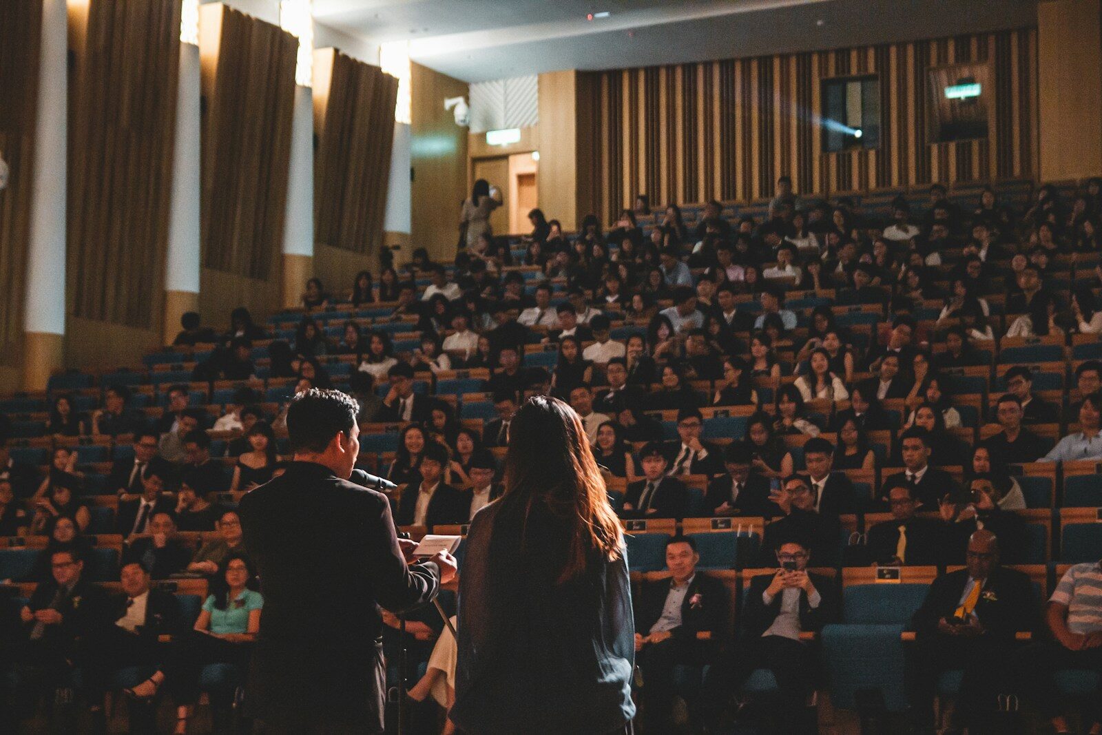 Two speakers address a large audience in an auditorium, emphasizing engagement and participation during the event. The crowd is attentive and diverse.