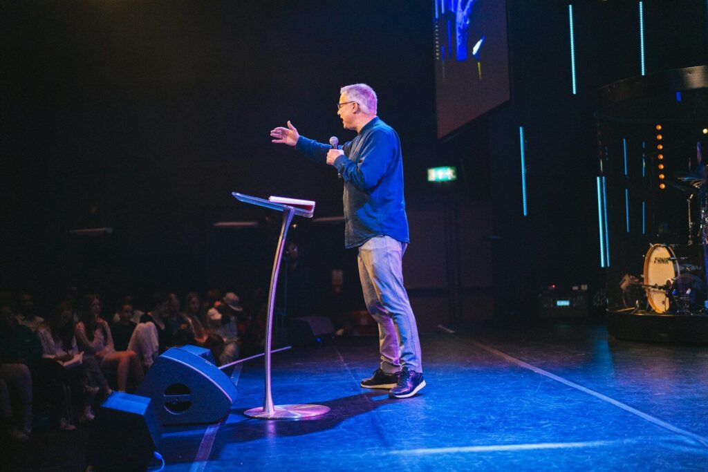 A speaker gestures animatedly at a podium during a presentation. The audience is engaged, seated in dim lighting with stage lights illuminating the scene.
